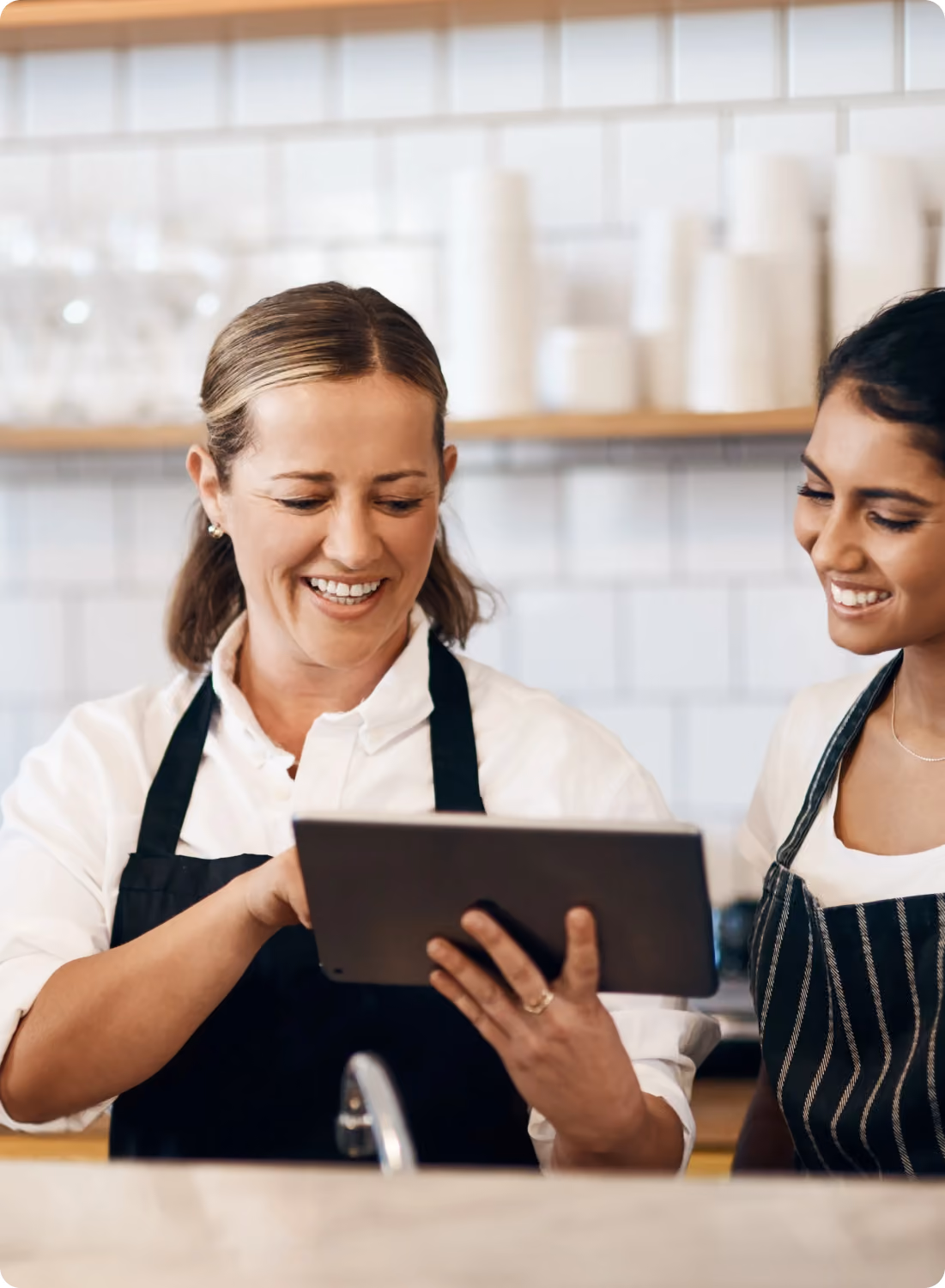 Two women in aprons smiling and looking at a tablet together in a kitchen setting with white tiled walls and shelves with cups behind them.