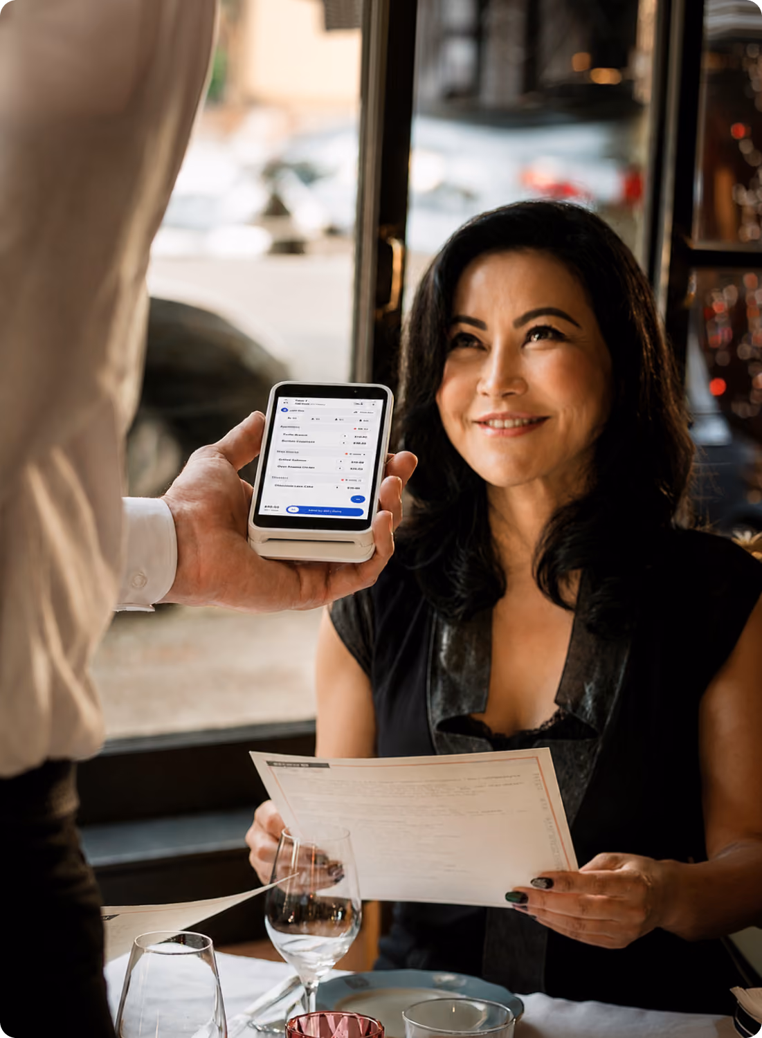 A woman sitting at a restaurant table smiling at a waiter who is holding a smartphone displaying a bill payment app, she holds a menu in her hands.
