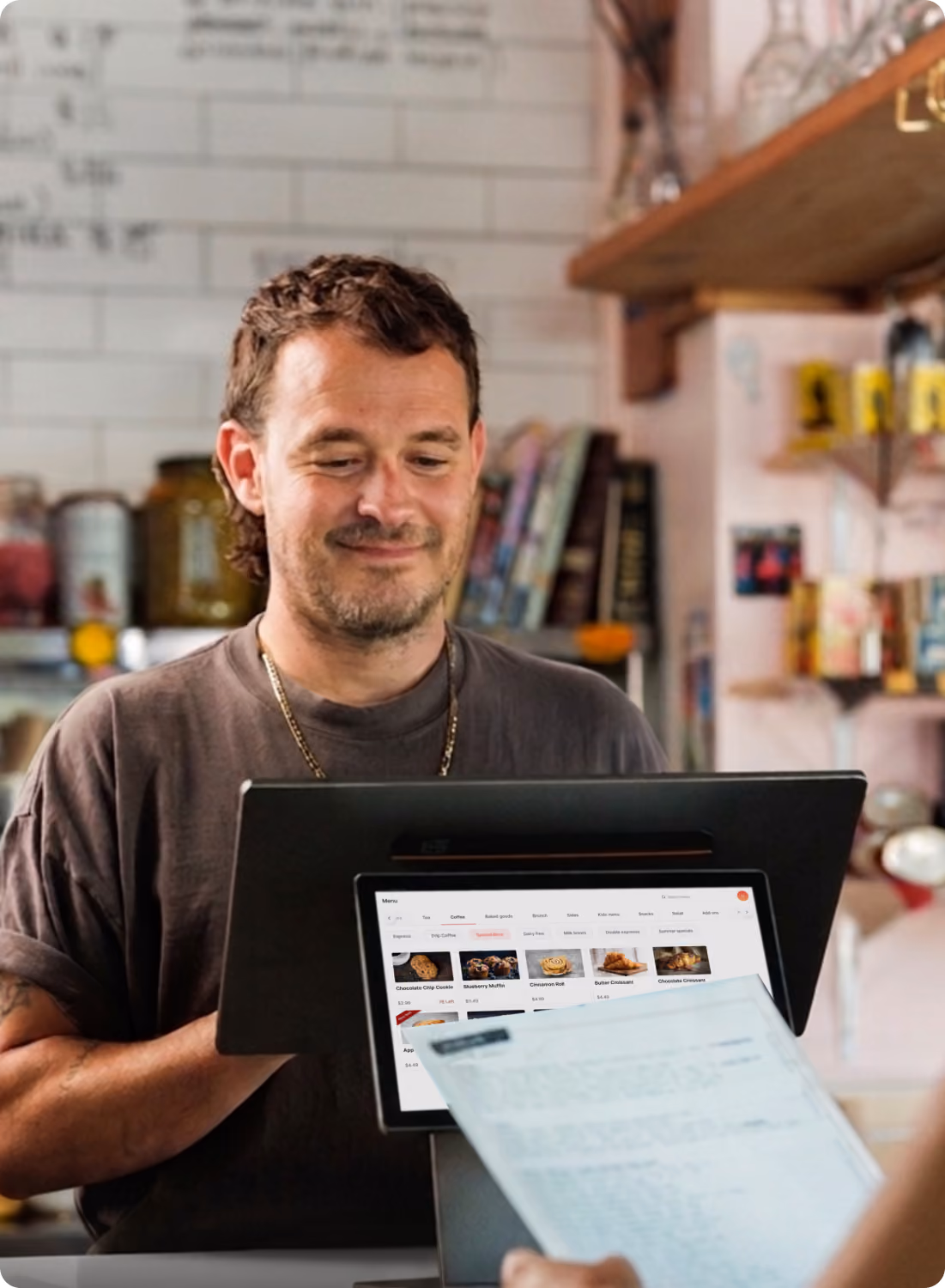 A smiling man with short brown hair and a short beard stands behind a black POS terminal displaying a digital menu with items like chocolate chip cookies and croissants, while a customer hands him a paper receipt in a cozy cafe setting.