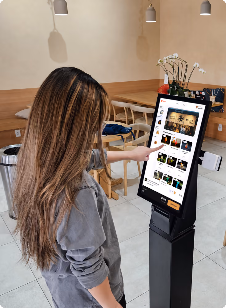 Woman with long hair wearing a gray shirt using a self-service touchscreen kiosk in a restaurant, selecting items from a digital menu on the screen.