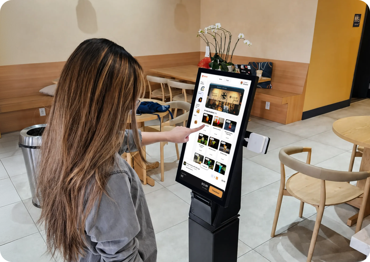 A woman with long hair selecting items on a touchscreen self-service kiosk inside a modern cafe with wooden chairs and tables, a trash can, and a flower decoration on the kiosk.