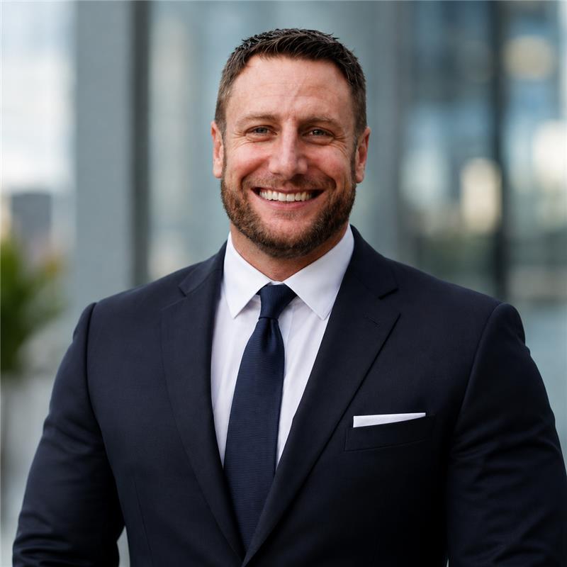 Smiling man with short brown hair and beard wearing a dark blue business suit, white shirt, and matching tie, standing in front of a blurred urban background.