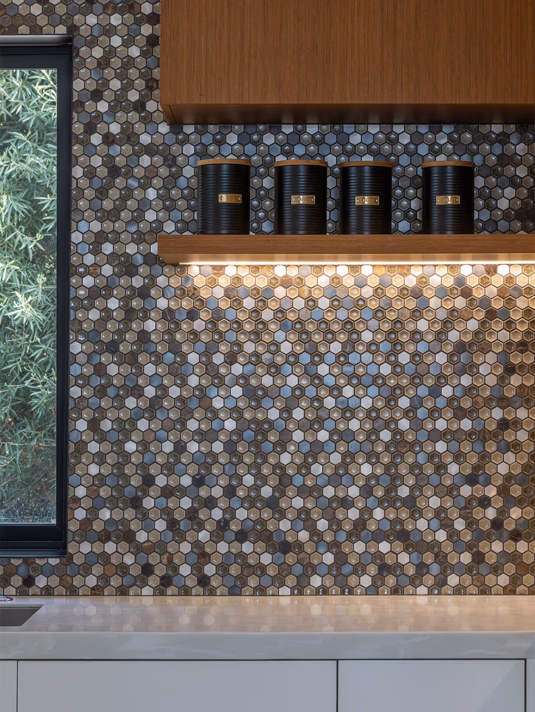 Kitchen backsplash with small hexagonal tiles in shades of gray and brown below wooden shelves with four black canisters labeled spices.