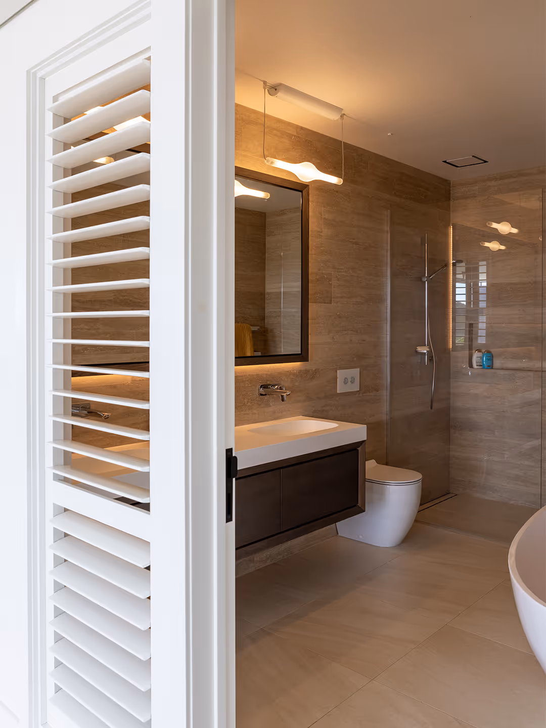 Modern bathroom with beige tiled walls, floating dark wood vanity with white sink, illuminated mirror, white toilet, and walk-in glass shower.