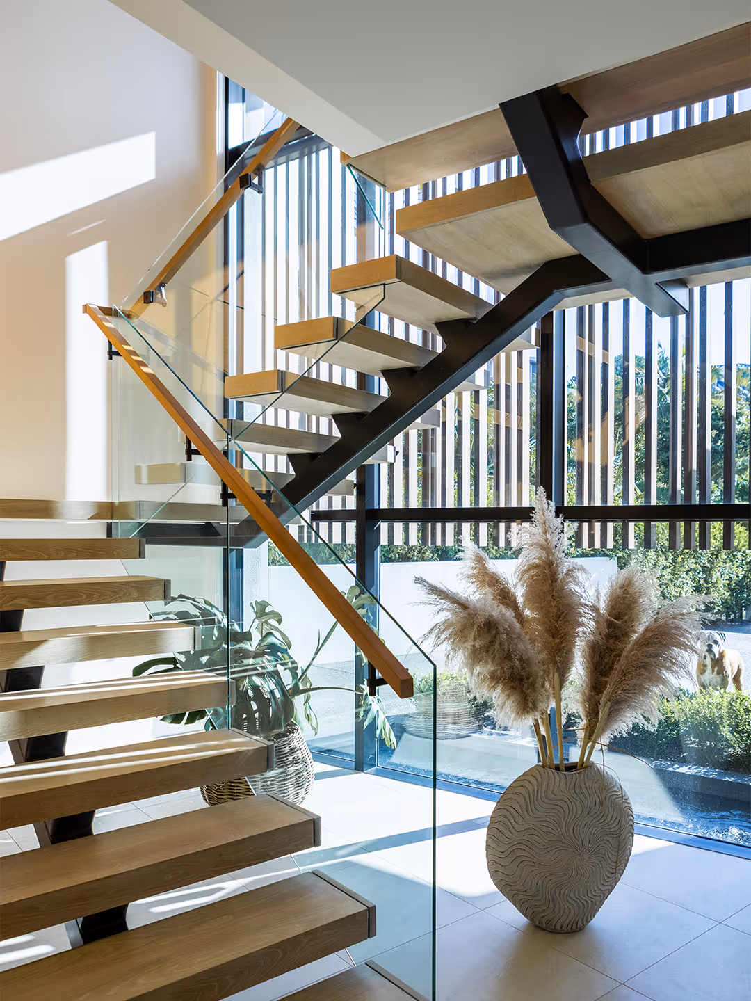 Modern wooden staircase with glass railing next to a vase with pampas grass in a sunlit room with large vertical windows.