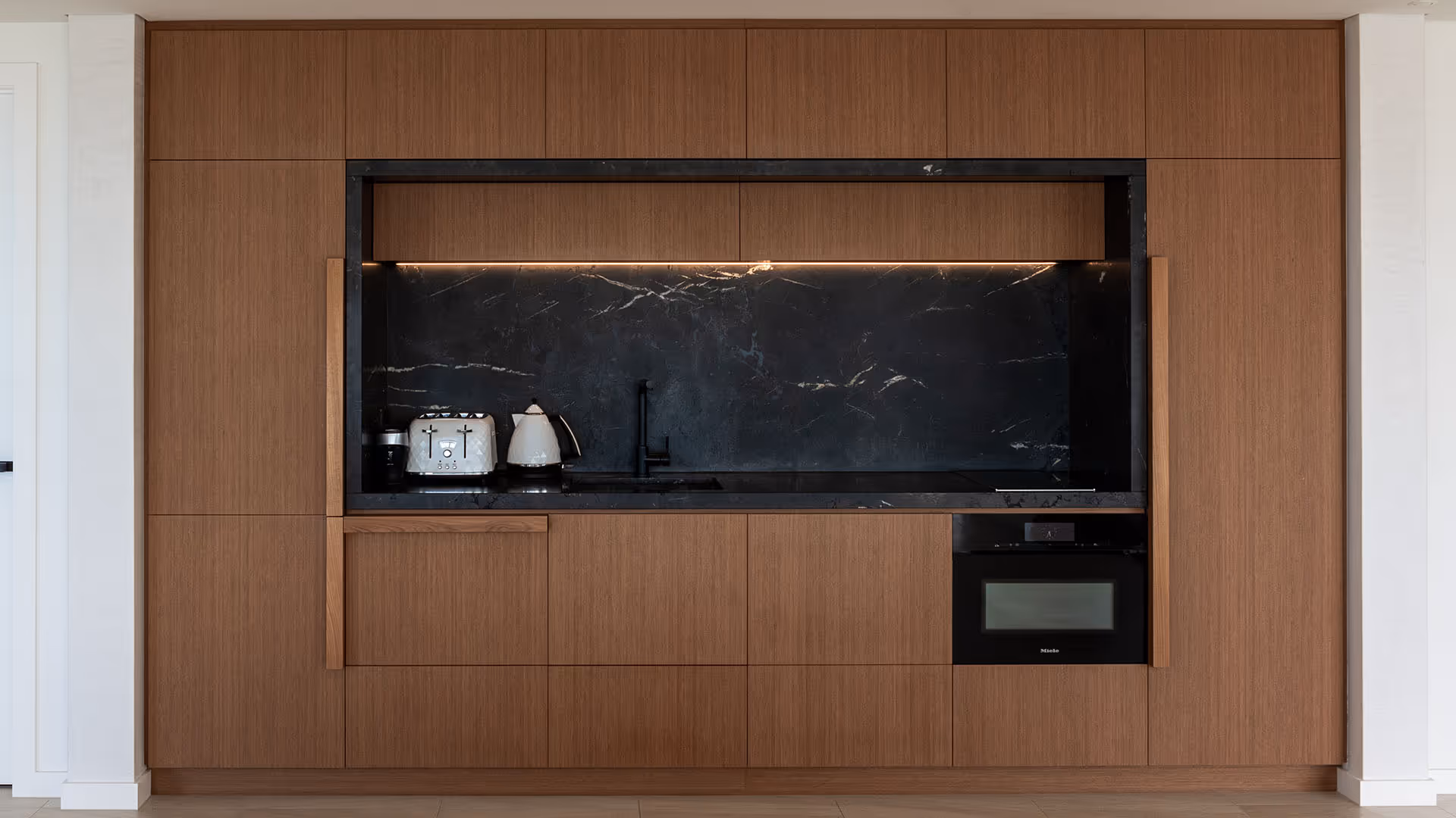 Modern kitchen wall with integrated wooden cabinets, black marble backsplash and countertop, white toaster and kettle, and built-in oven.
