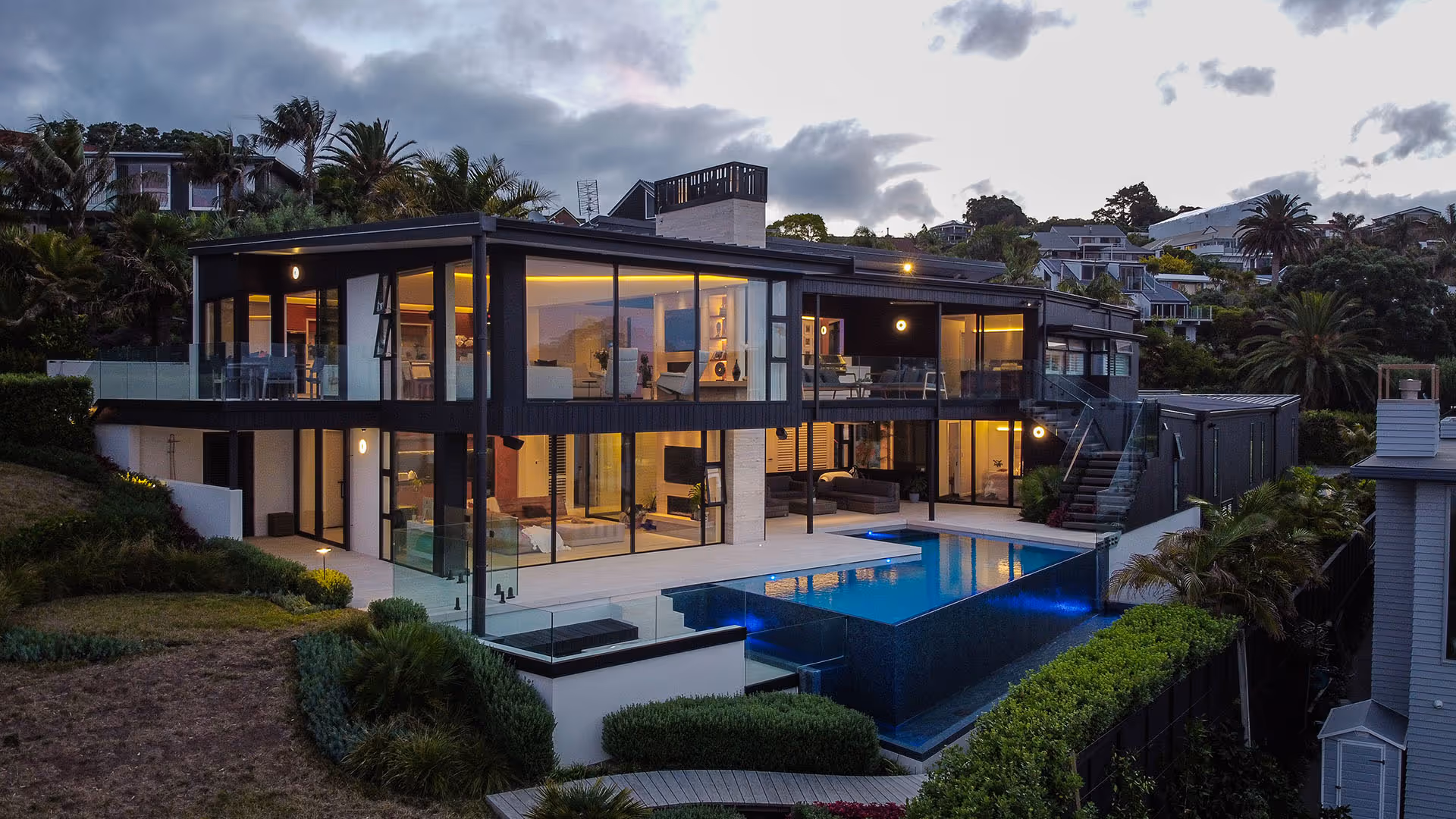 Modern two-story house with large glass windows and a lit interior overlooking an illuminated infinity pool at dusk.