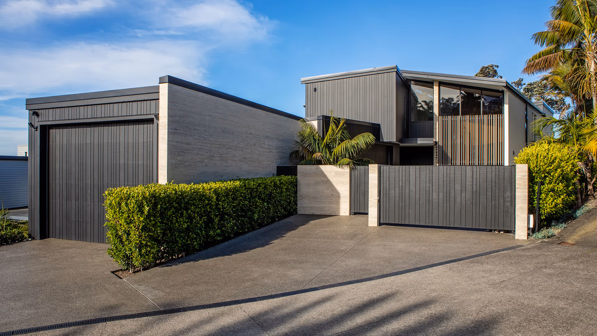 Modern house with dark vertical paneling, large windows, a gated driveway, and lush green hedges under a blue sky.