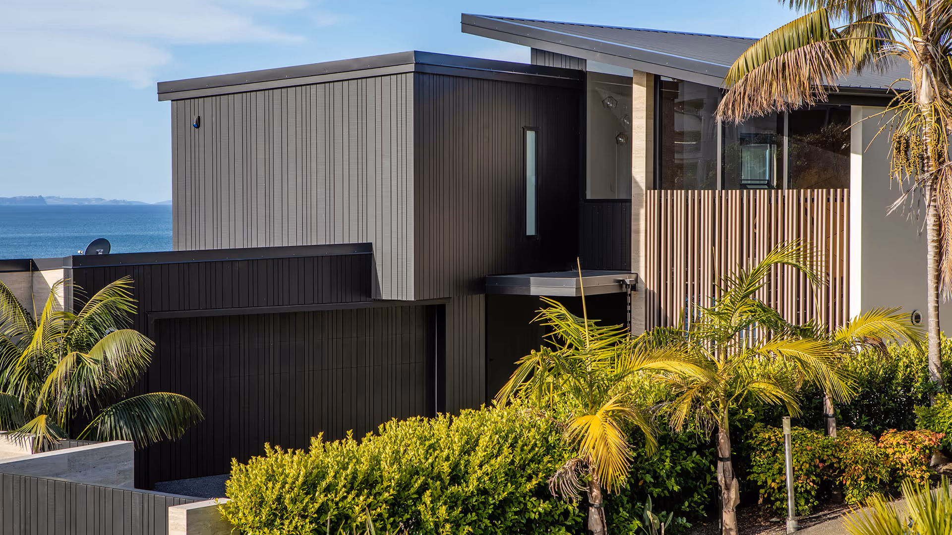 Modern black and gray two-story house with vertical siding, surrounded by palm trees and greenery with ocean view in the background.