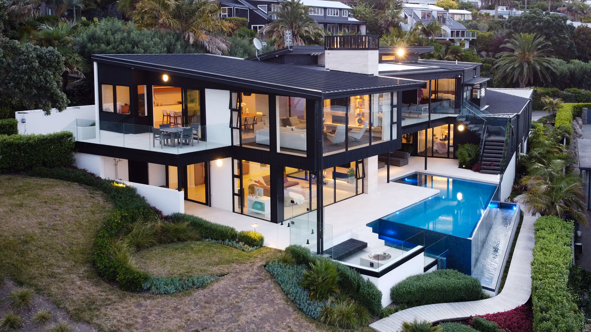 Modern two-story house with large windows, illuminated interior, balcony, and an infinity pool surrounded by landscaped greenery.