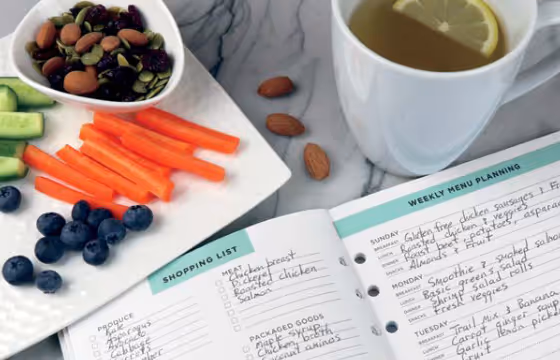 Plate with mixed nuts, carrot sticks, blueberries, cucumber slices next to a cup of lemon water and a handwritten weekly menu planner and shopping list on a marble surface.