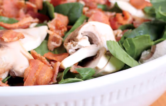 Close-up of a salad with fresh spinach leaves, sliced mushrooms, and crispy bacon bits in a white bowl.