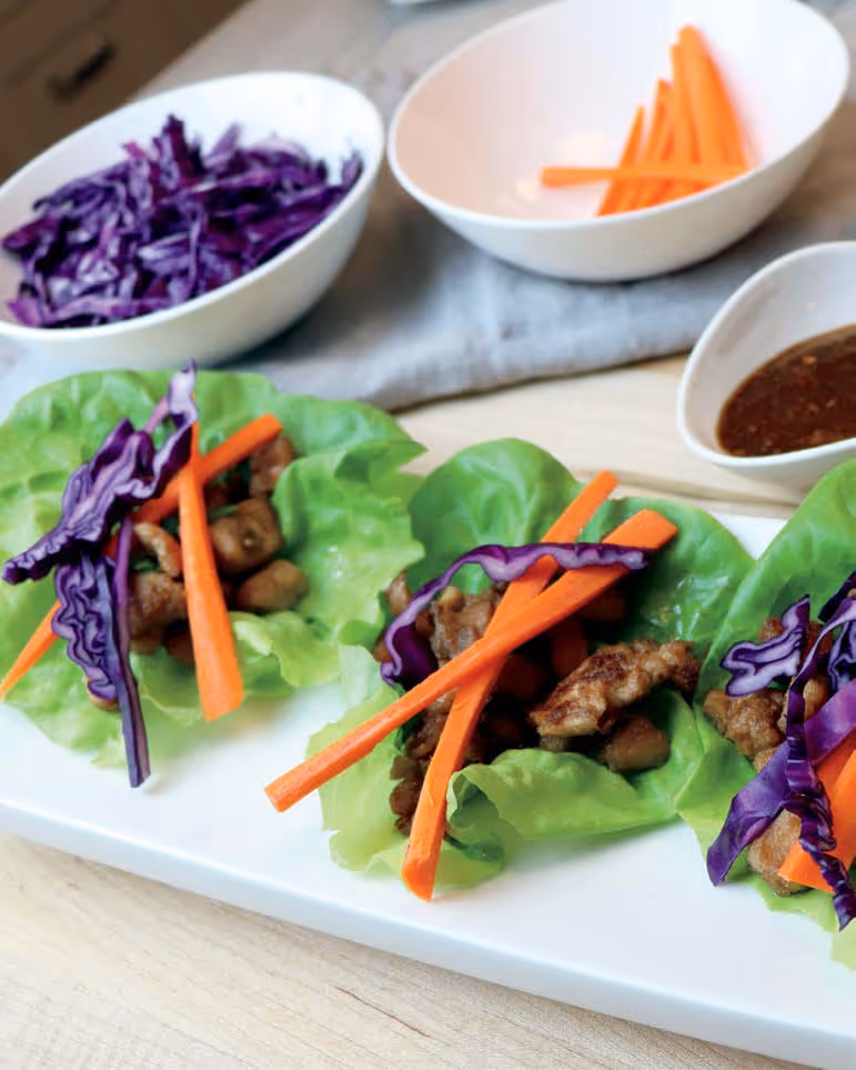 Three lettuce wraps filled with cooked meat, shredded purple cabbage, and carrot sticks on a white rectangular plate with bowls of shredded cabbage, carrot sticks, and dipping sauce in the background.