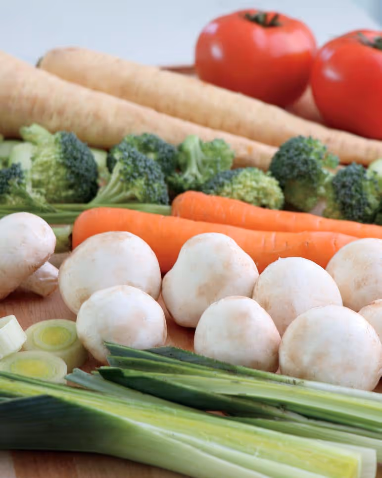 Assorted fresh vegetables including white mushrooms, carrots, broccoli, tomatoes, parsnips, and leeks on a wooden surface.