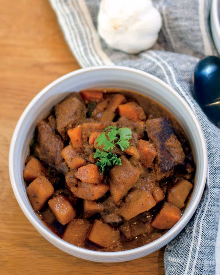 Bowl of beef stew with chunks of meat and sweet potatoes garnished with fresh parsley on a wooden table.