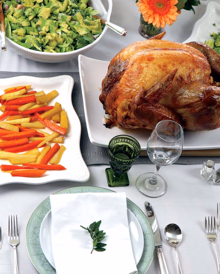 Roast chicken on a white platter, bowl of mixed green salad with avocado, plate of roasted carrot and parsnip sticks, table set with green-patterned plates, silver cutlery, and green and clear glasses.