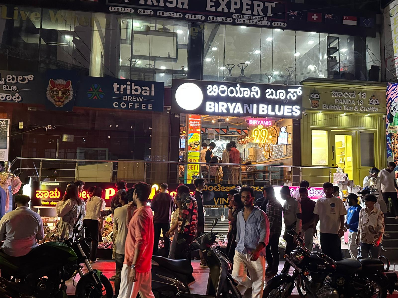 インド・バンガロールの夜の繁華街。明るくライトアップされた飲食店が並び、『BIRYANI BLUES』『PANDA 18 Bubble Tea & Croffle』『tribal brew COFFEE』などの店舗名が英語とカンナダ語で表示されている。人々がバイクや歩行でにぎやかに通りを行き交い、BIRYANI BLUESの店内には多くの客が見える。路面店の看板にある言語は英語とカンナダ語。