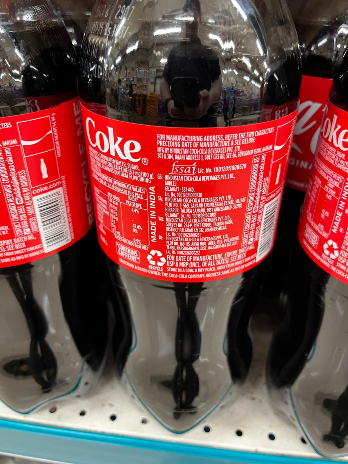 Coca-Colaの1.25リットルペットボトルの裏面ラベル。赤地に白い英語表記で、原材料（炭酸水、砂糖、カラメル色素など）、カフェイン含有、栄養成分表示、製造業者名（Hindustan Coca-Cola Beverages）、製造地（インド）、再利用マーク、使用上の注意などが記載されている。