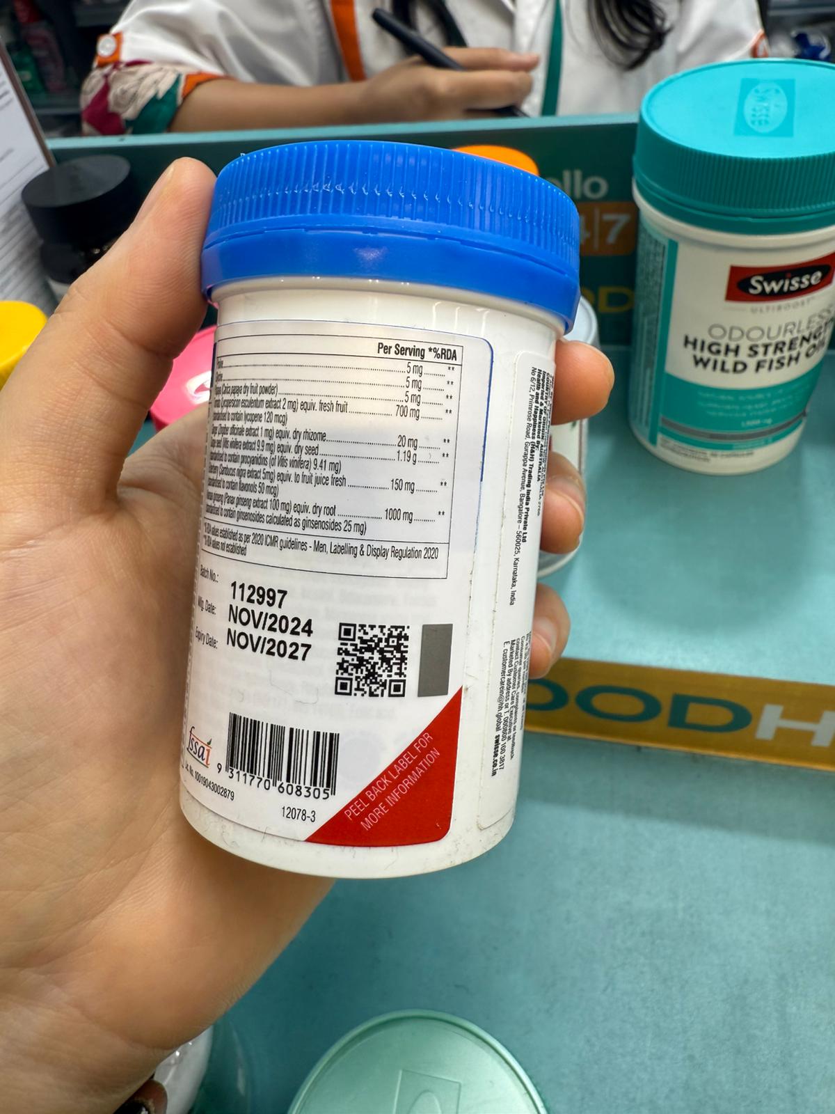 白いプラスチック容器に青いフタが付いた健康補助食品の側面ラベル。英語で1回分あたりの栄養成分（ビタミンや植物エキスなど）が細かく記載され、下部には製造ロット番号、有効期限（2024年11月製造、2027年11月まで）、QRコード、バーコード、FSSAIマーク、『詳細情報はラベルをめくって確認』という案内がある。記載言語は英語のみ。