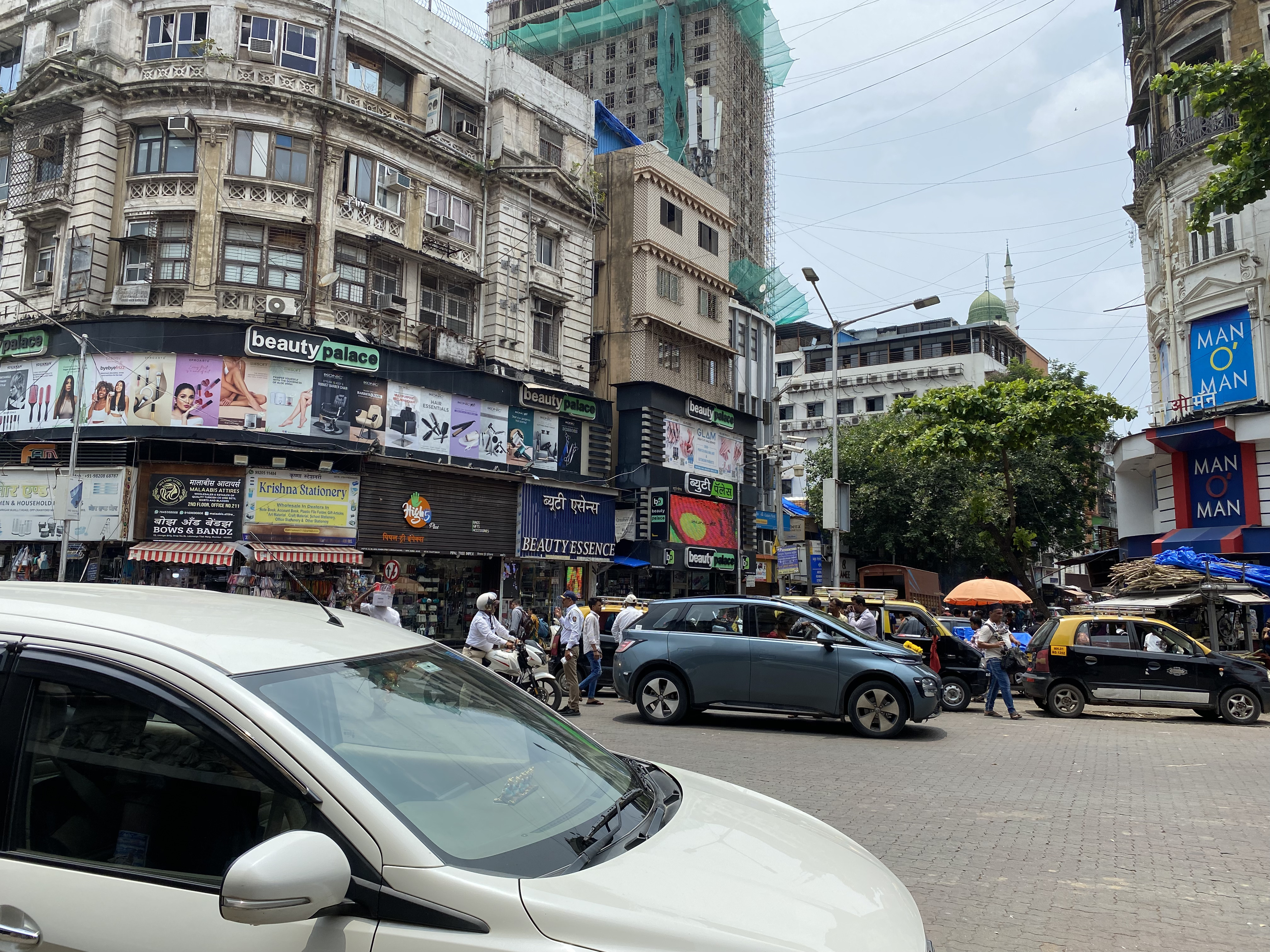 インド・ムンバイの市街地の交差点。古い西洋風の建物が並び、1階部分には『beauty palace』や『Krishna Stationery』『MAN O’ MAN』など英語とヒンディー語の看板が多数掲げられている。タクシーや自家用車、バイクが行き交い、歩行者や商店の人々でにぎわっている。遠景にはモスクの尖塔も見える。路面店の看板にある言語は英語とヒンディー語。