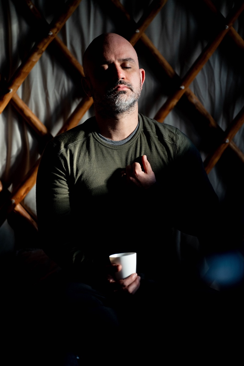 A man sits with eyes closed holding a cup in a shaft of light inside the yurt at The Return of Men retreat, 2024.