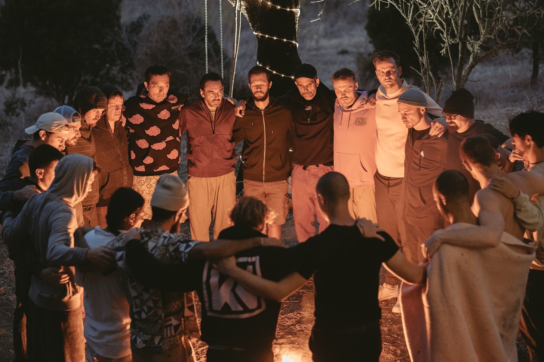 The circle stands arm over shoulder at dusk beneath a lit tree at The Return of Men retreat, October 2025.