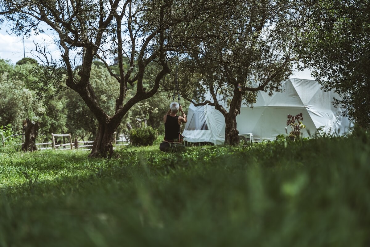 A man with grey hair sitting alone on a swing beneath olive trees at a men's retreat in Arrábida, Portugal, in a quiet moment of reflection