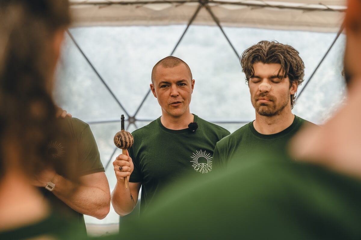 A facilitator raises a rattle as the men stand with closed eyes at The Return of Men retreat, Arrábida, 2026.