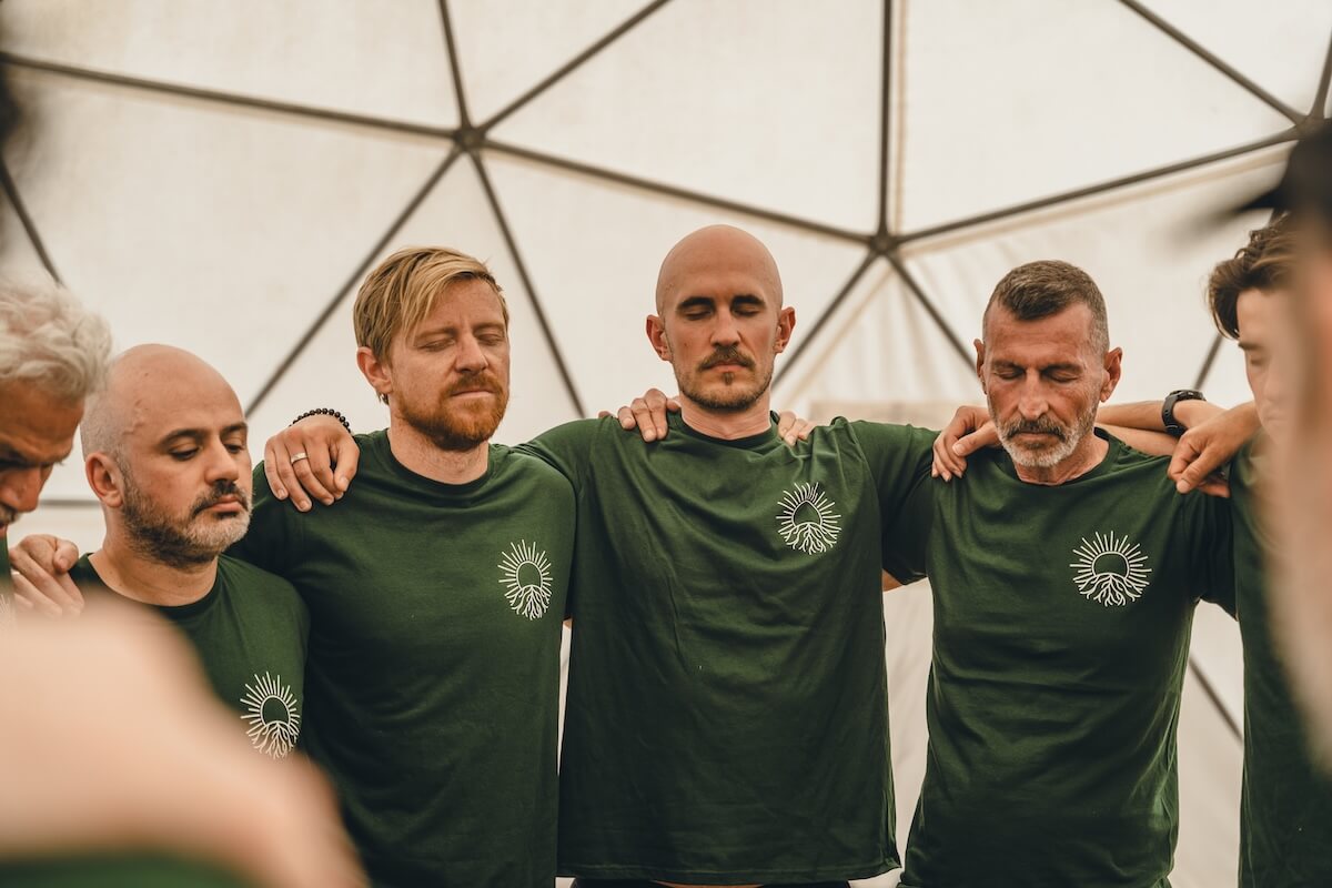 Men stand arm in arm with eyes closed in their retreat shirts at The Return of Men, BioVilla, May 2026.