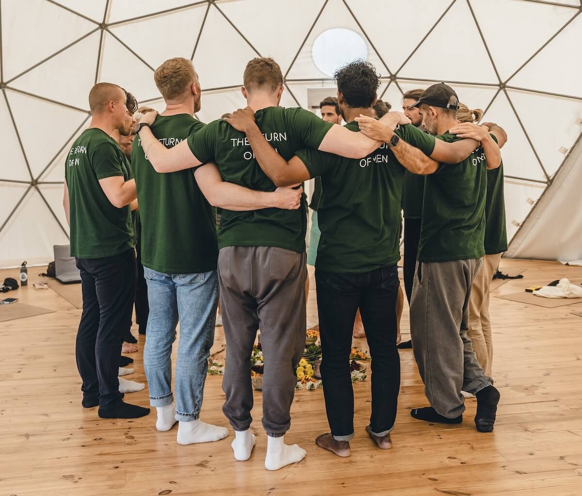 The men huddle arm in arm around the flower mandala at the close of The Return of Men retreat, May 2026.