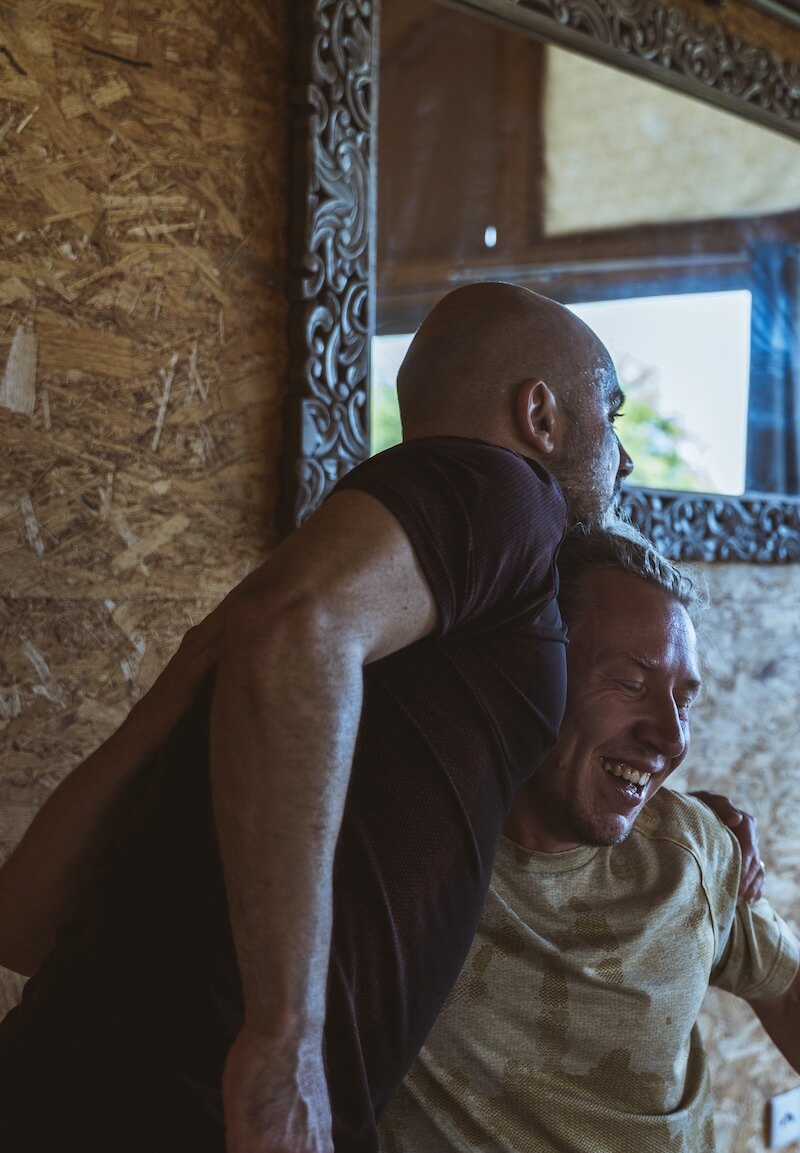 Two men laugh in a close embrace by the mirror at The Return of Men retreat, Arrábida, Portugal, 2026.