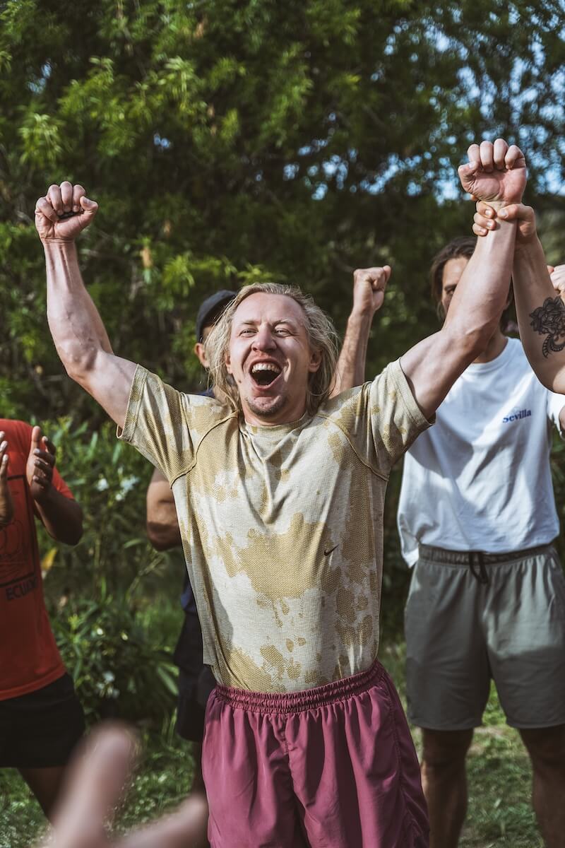 A soaked man roars in triumph, arms raised, as the others cheer at The Return of Men retreat, May 2026.