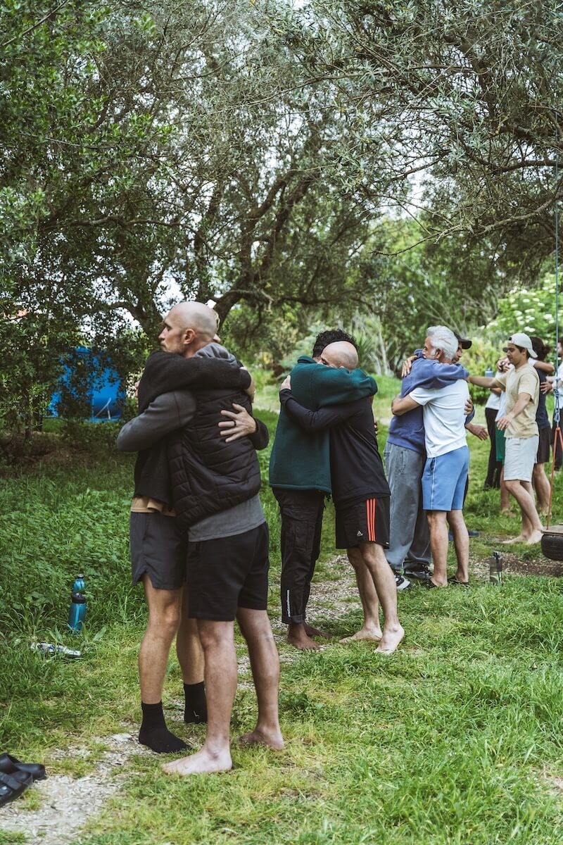 The men embrace in pairs beneath the olive trees at The Return of Men retreat, Arrábida, Portugal, May 2026.