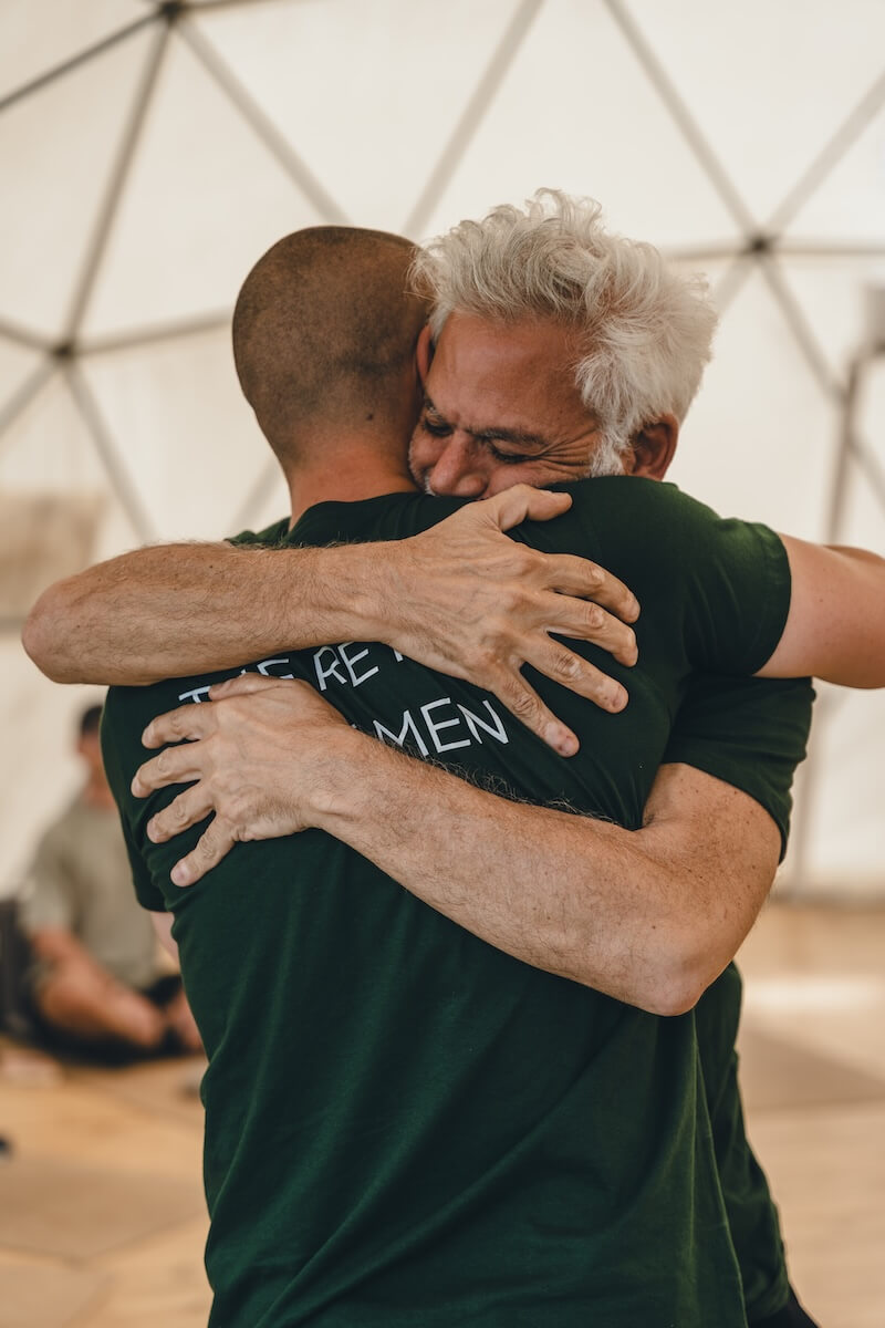 A silver-haired man is held through tears in a long embrace at The Return of Men retreat, BioVilla, May 2026.