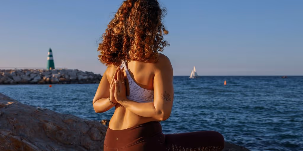 woman sitting while doing yoga on rock viewing body of water during daytime