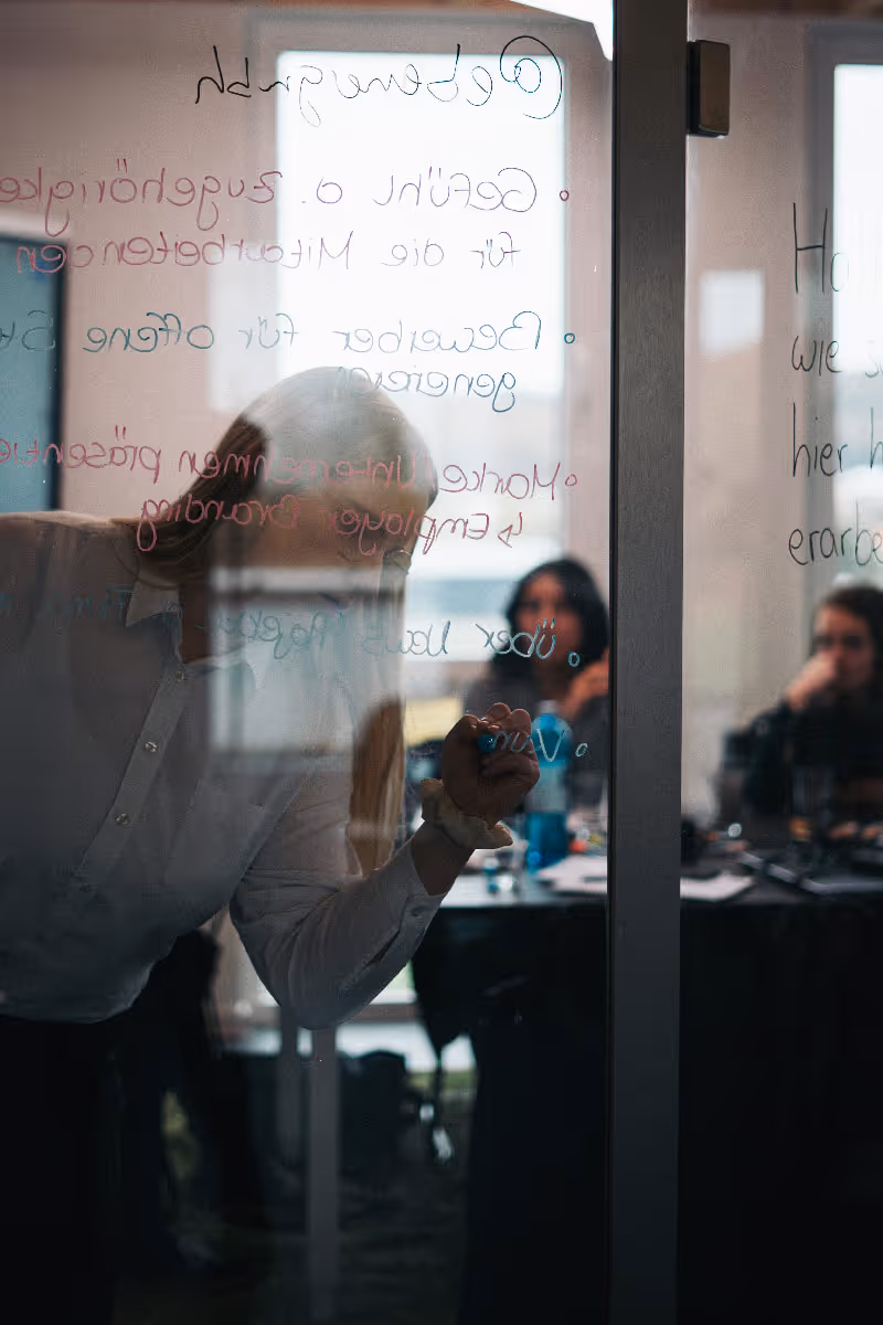 Frau schreibt mit Marker etwas auf eine Glasscheibe. Zwei Frauen im Hintergrund schauen zu. Social Media Workshop
