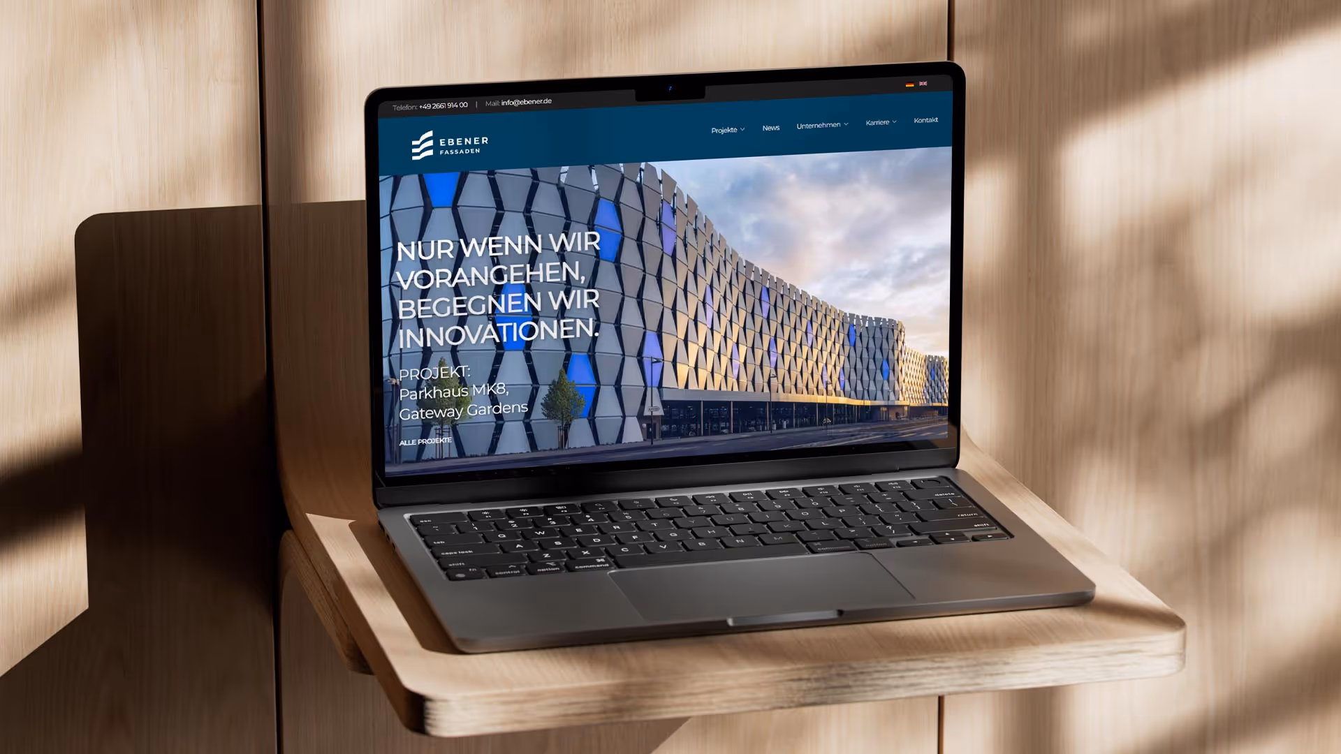 Laptop auf einem Holzständer vor einer Holzwand. Aufgeklappt und zeigt eine Website.
