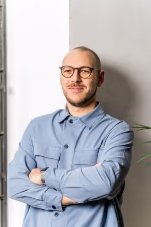 Mann mit Brille und blauem Hemd lehnt mit verschränkten Armen an einer hellen Wand und lächelt.
