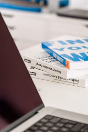 Drei gestapelte Bücher über Design und Geschäftsmodelle neben einem geöffneten Laptop auf einem weißen Schreibtisch.