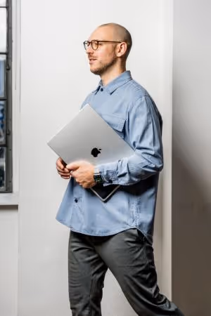 Mann mit Brille, der ein geschlossenes MacBook in den Händen hält und an einer weißen Wand steht.
