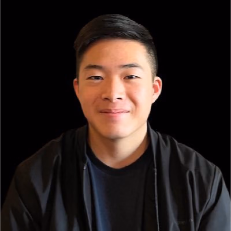 Young Asian man with short black hair, wearing a black jacket and black shirt, smiling slightly.