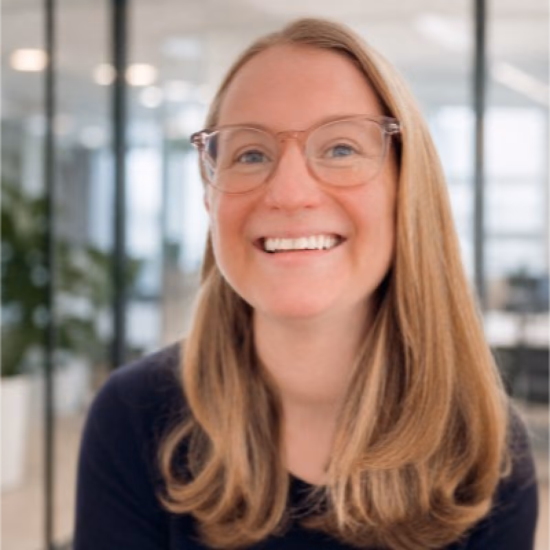 Smiling woman with long light brown hair wearing glasses and a dark top in a bright office setting with glass partitions and plants.