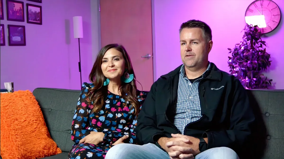 A couple sitting in a purple room on a gray couch with orange pillow