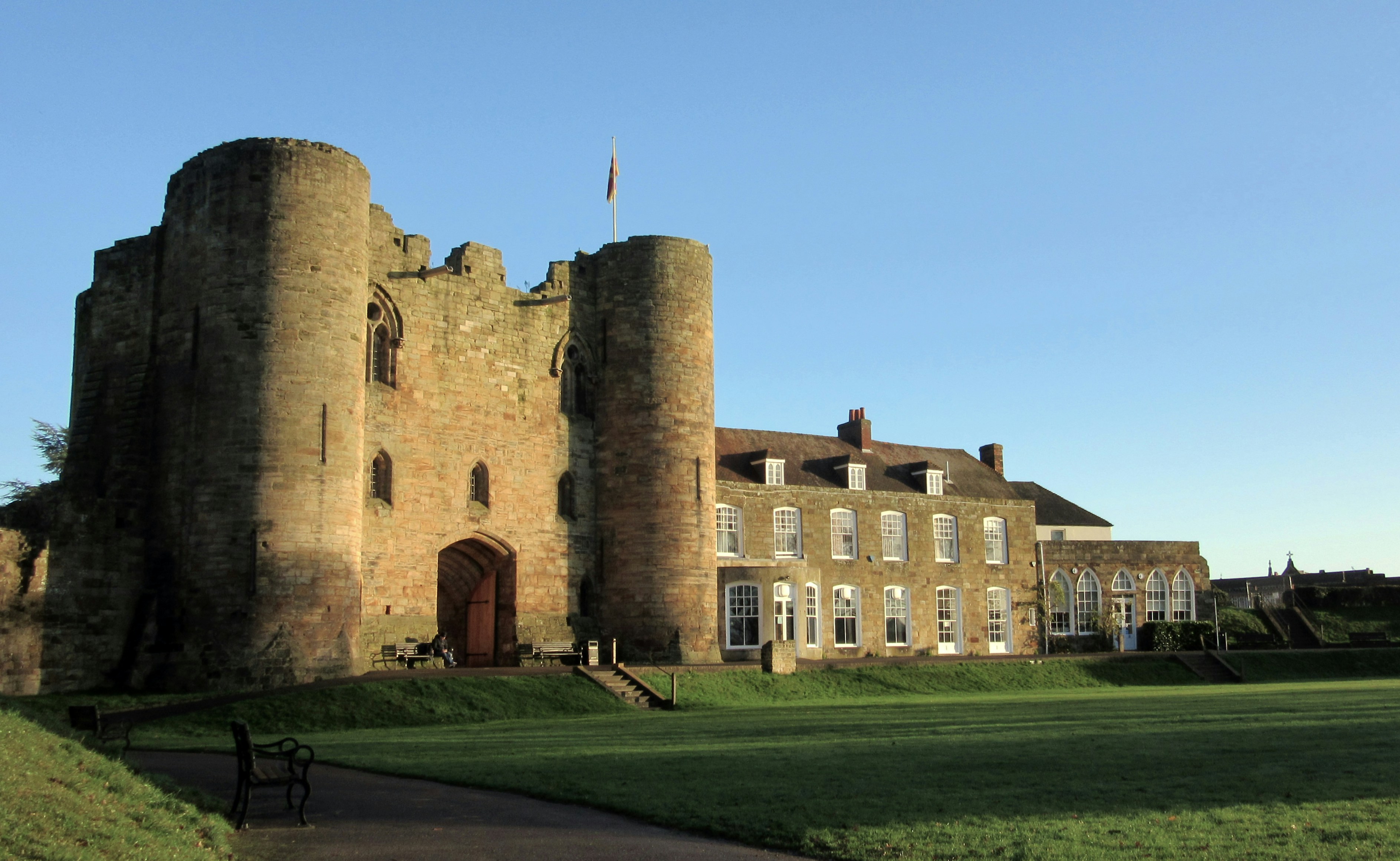 Historic stone castle with round towers and adjoining building under clear blue sky, with green lawn in front.