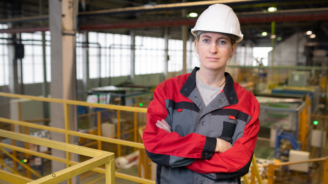 Woman in a red and gray work jacket and white hard hat standing with arms crossed in an industrial factory setting.