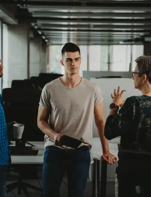 A man in a gray t-shirt holding a tablet, standing in an office setting next to a person gesturing with their hands, representing flexible hiring models offer