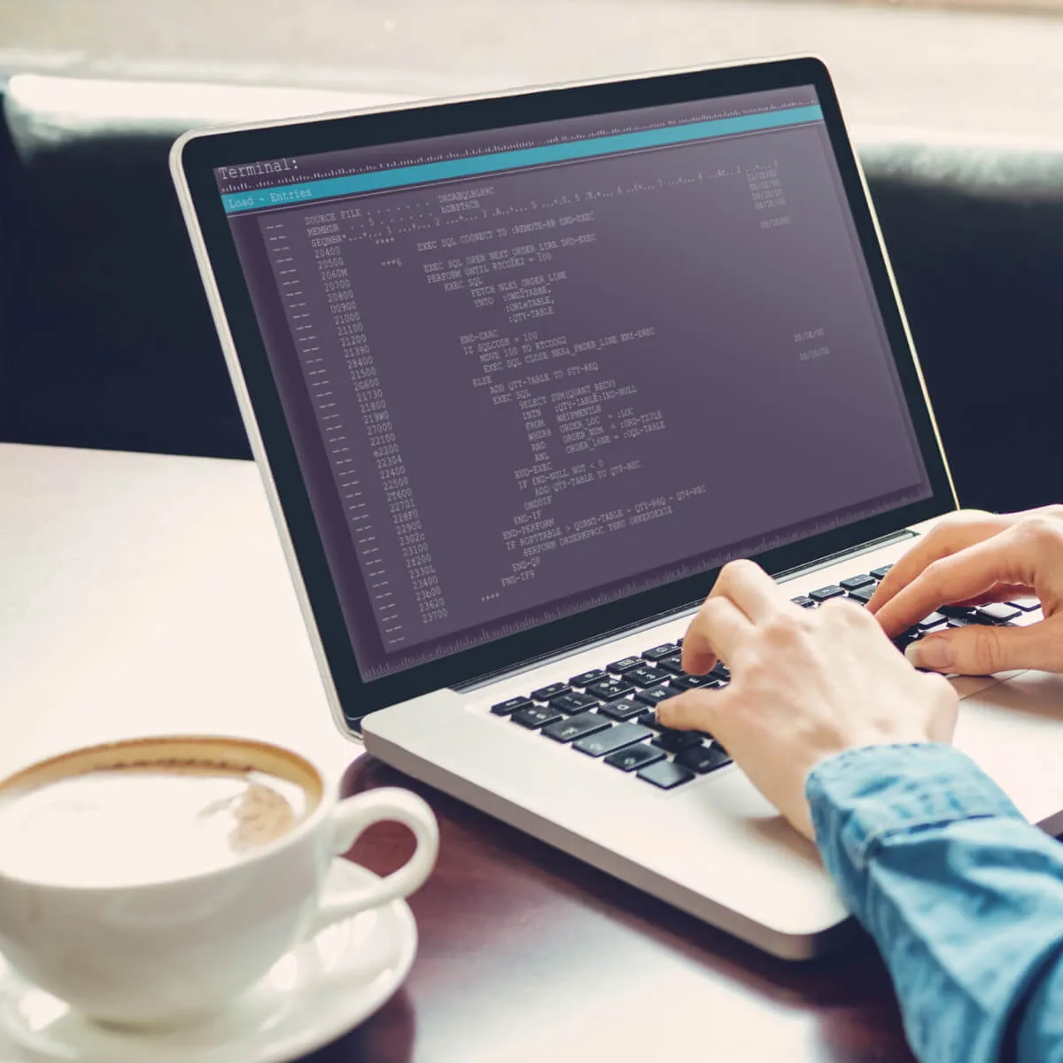 The image shows a person typing on a laptop keyboard. A terminal window with a code is displayed on the laptop screen. The code appears to be written in a programming language, possibly related to Job Control Language (JCL) or a similar scripting language used for batch processing on mainframes. The terminal window has a title bar that says "Terminal" and a menu bar with options like "Load" and "Entities". There is also a cup of coffee on the table next to the laptop.