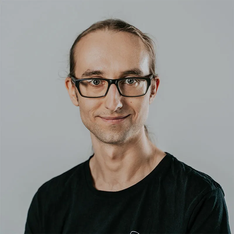 Portrait of Tomasz Swistak, Software Architect at Synergy Codes with long, light brown hair tied back, wearing black rectangular glasses and a black T-shirt.