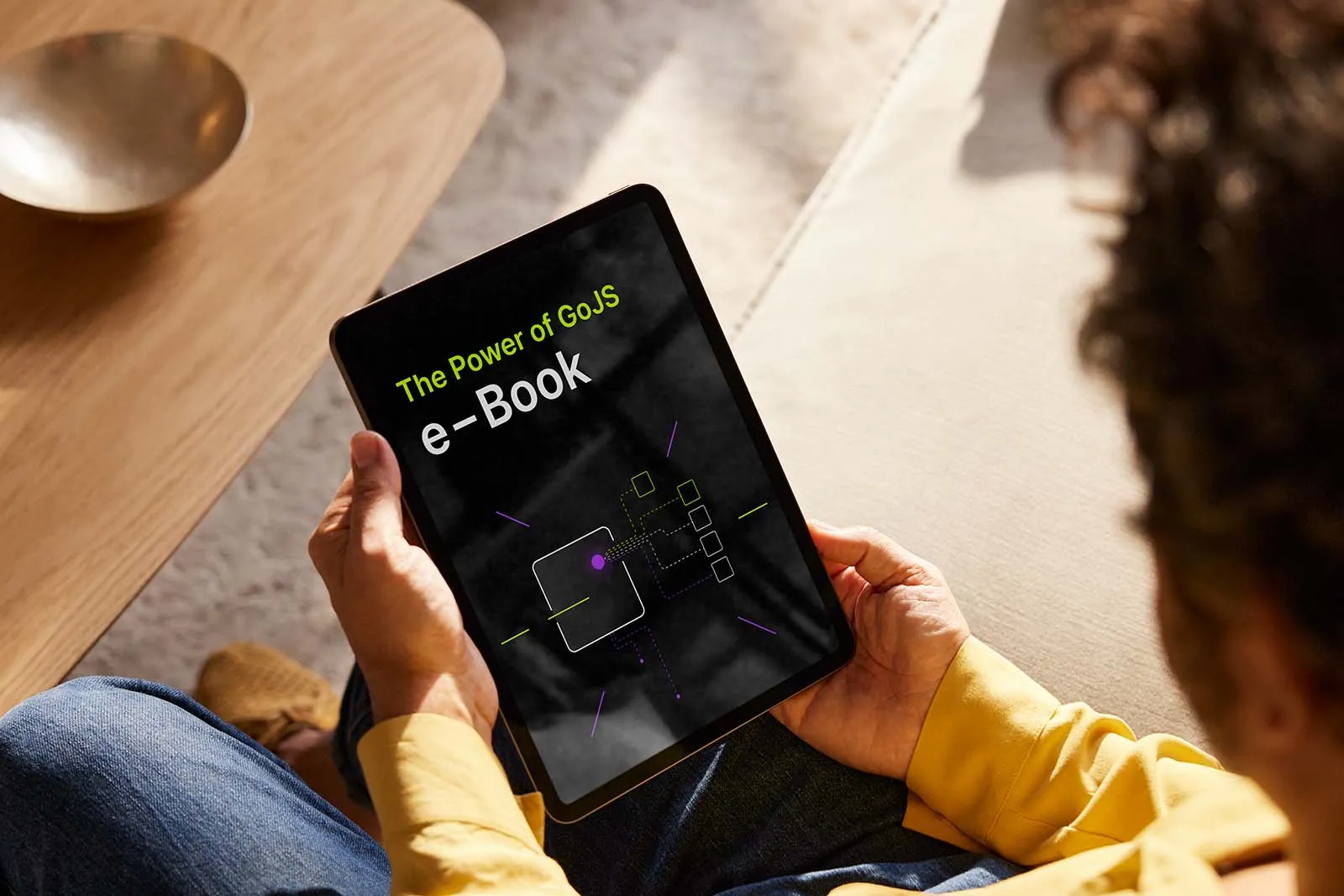 A person holding a tablet with the cover of a GoJS ebook titled ‘The Power of GoJS’ is shown. The eBook cover features abstract data visualization elements and a modern, minimalist design with purple highlights. The person is sitting on a couch, wearing a yellow shirt and jeans, with a wooden coffee table and a decorative bowl in the background.