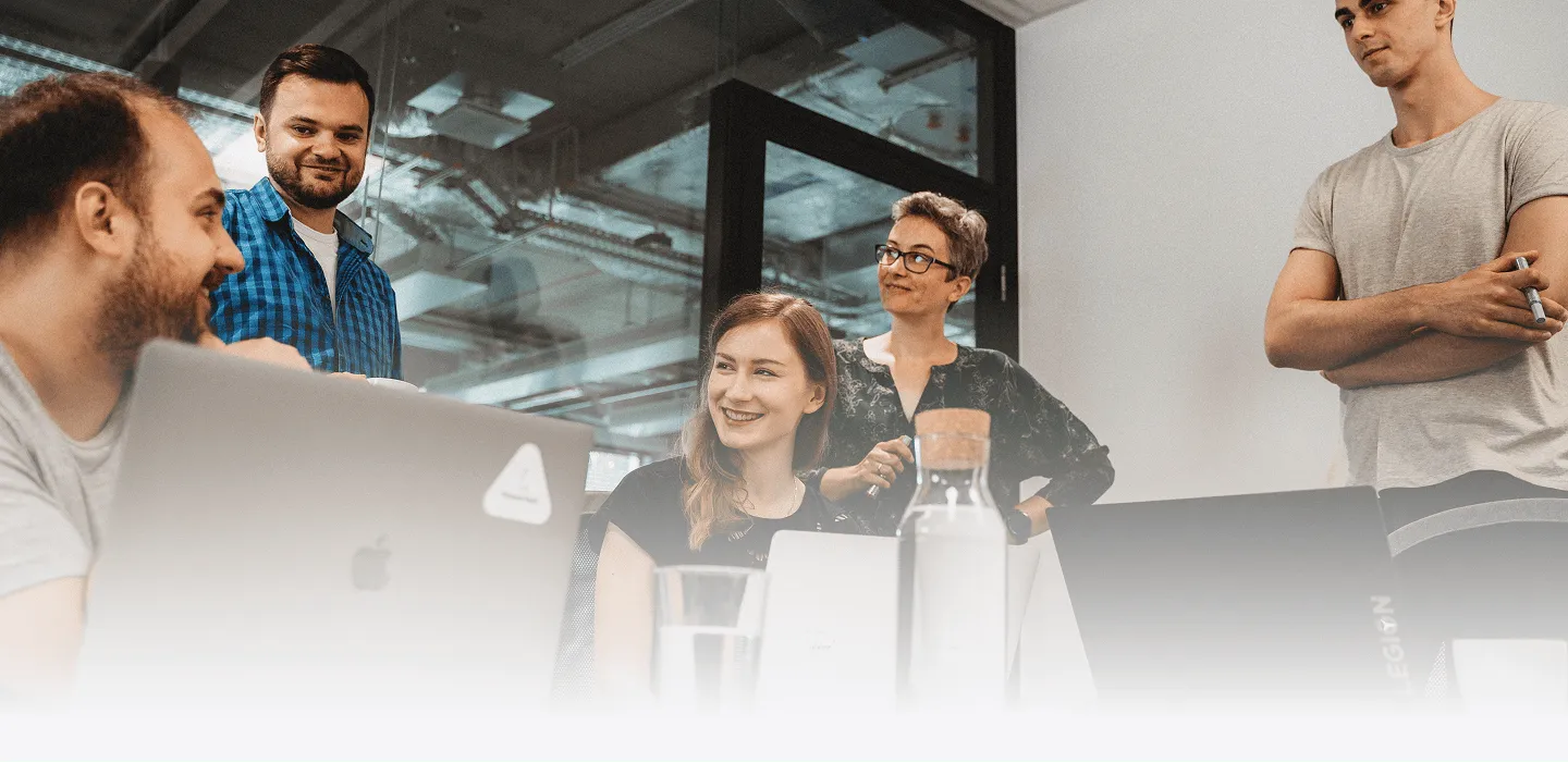 A group of team members gathered around a table during a collaborative meeting in a modern office, smiling and working on laptops.